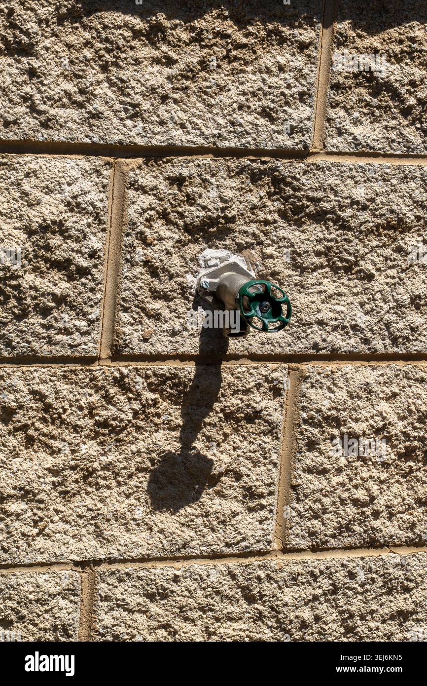 Un mur en pierre texturé avec un robinet d'eau extérieur unique avec un tuyau métallique et une poignée verte circulaire Banque D'Images