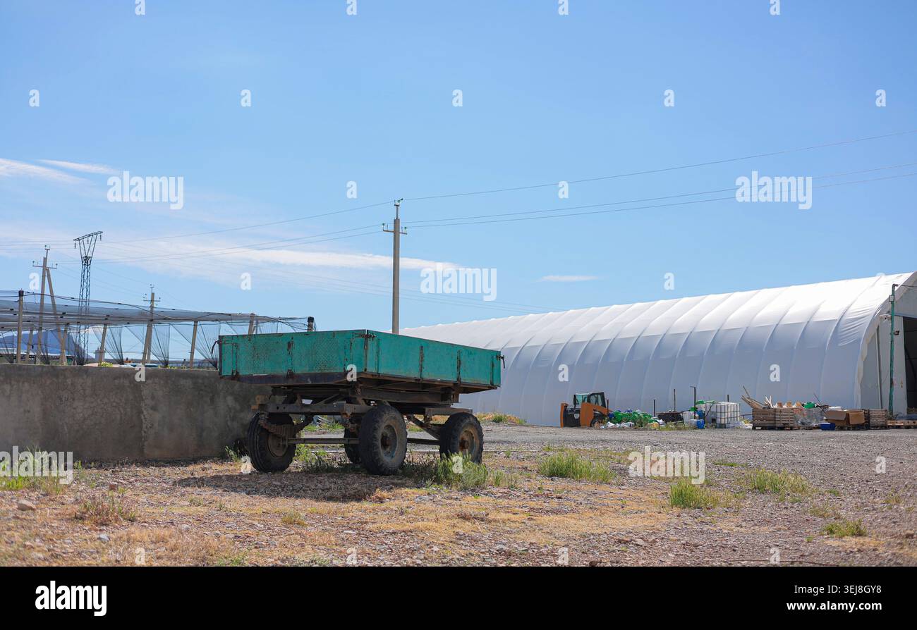 Une ancienne remorque agricole verte garée dans une cour de ferme devant un ciel bleu et un entrepôt blanc. Banque D'Images