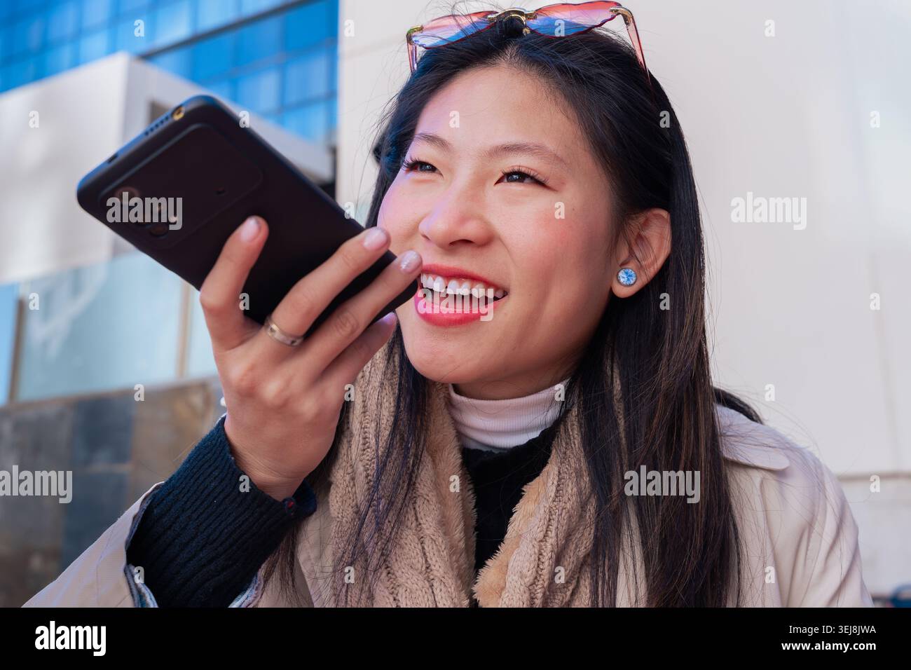 Jeune femme asiatique tenant le téléphone envoyant une note vocale et souriant en dehors de la zone urbaine Banque D'Images