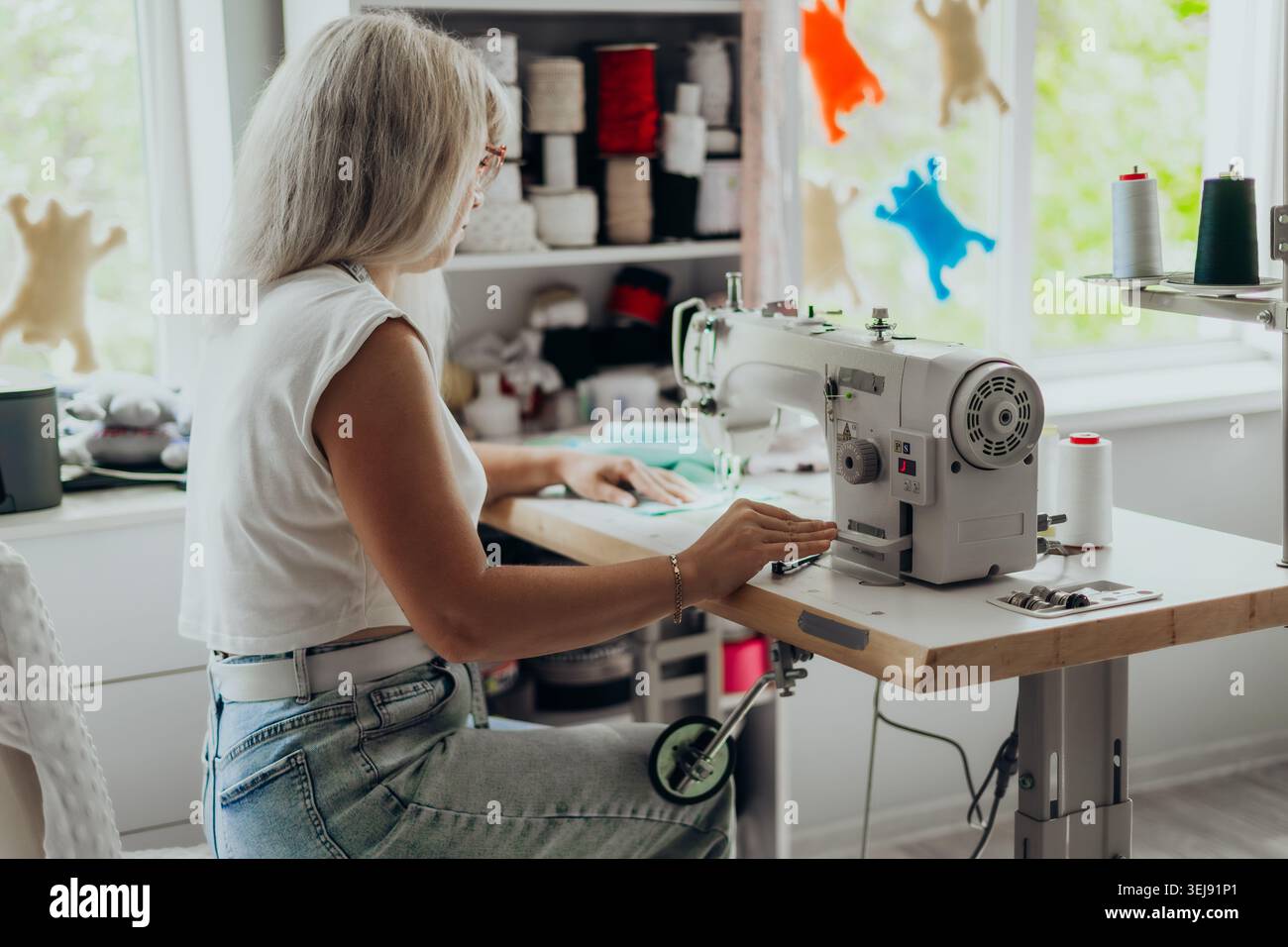 Femme avec du tissu à coudre de cheveux gris sur une machine à coudre professionnelle dans un studio moderne lumineux. Espace de travail créatif avec des fils colorés et fait à la main Banque D'Images