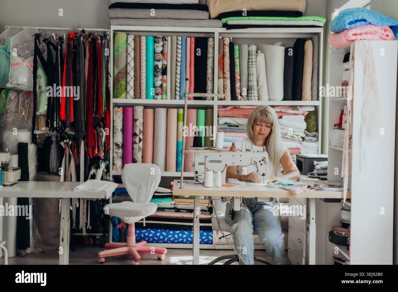 Créateur de mode travaillant dans un studio créatif rempli de tissus colorés et de matériaux de couture. Femme cousant à une machine professionnelle, entourée par Banque D'Images