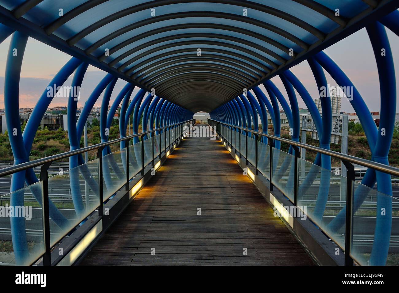 Pont piétonnier couvert bleu avec de fortes lignes de perspective et une passerelle éclairée au-dessus d'une route urbaine au crépuscule. Banque D'Images