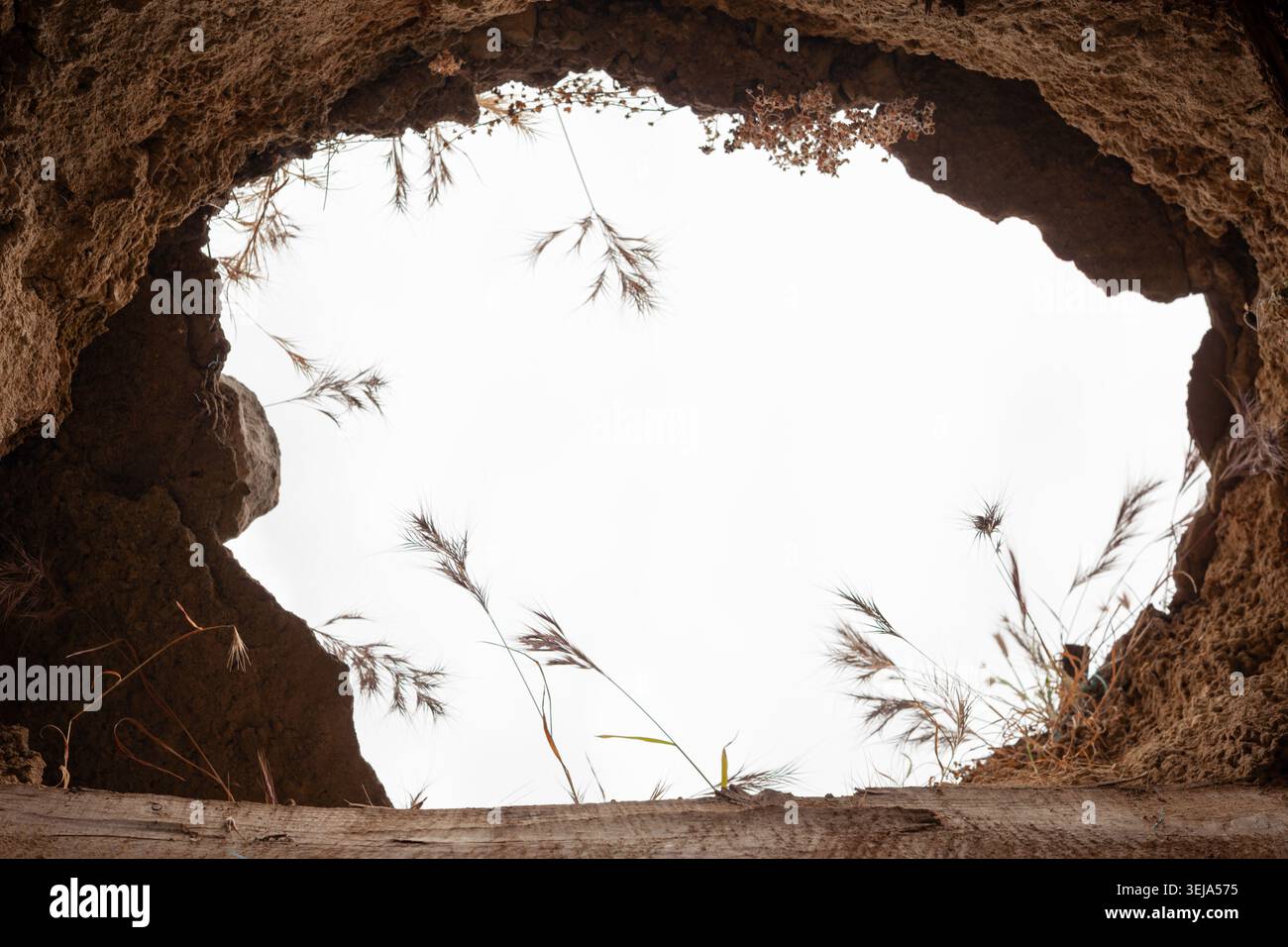 Une perspective de bas en haut depuis l'intérieur d'un trou sombre dans la terre ou d'une grotte souterraine révèle une ouverture circulaire encadrée par de la terre rugueuse et de l'herbe sèche. Banque D'Images