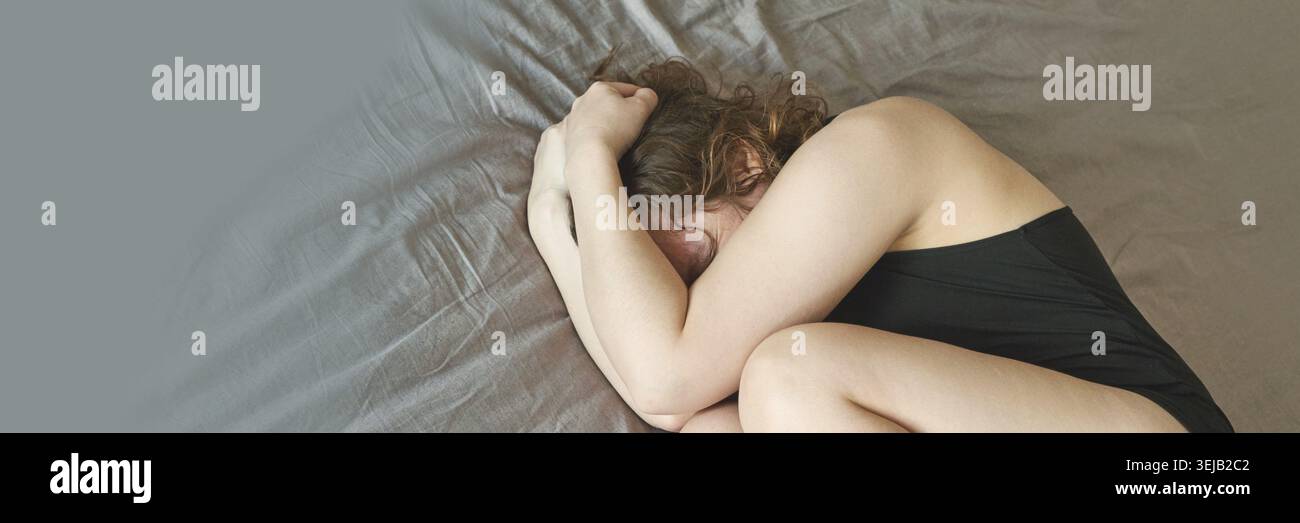 Jeune femme caucasienne en vêtements noirs enroulée en position fœtale sur le lit avec des draps gris montrant la détresse émotionnelle Banque D'Images