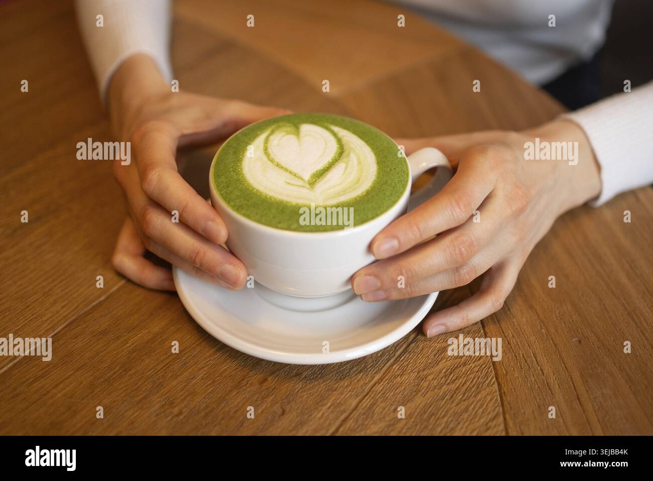 Mains de femme tenant une tasse blanche de latte matcha décorée d'un art latte en forme de cœur, placée sur une table en bois, créant un atmo confortable et invitant Banque D'Images