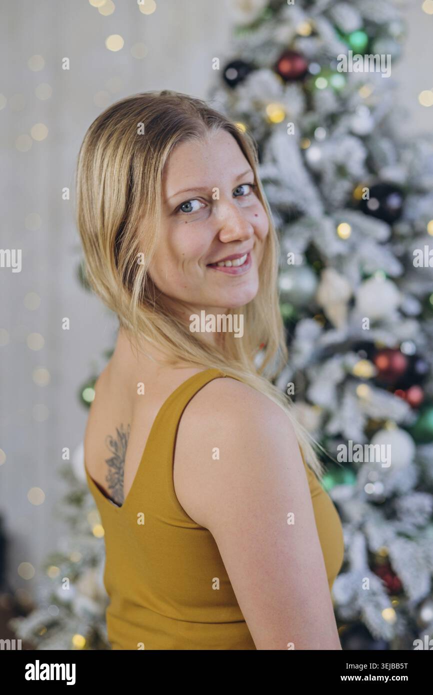 Jeune femme blonde avec tatouage sur son dos souriant à côté de l'arbre de noël décoré avec des lumières de fée et des boules colorées pendant les vacances d'hiver c Banque D'Images