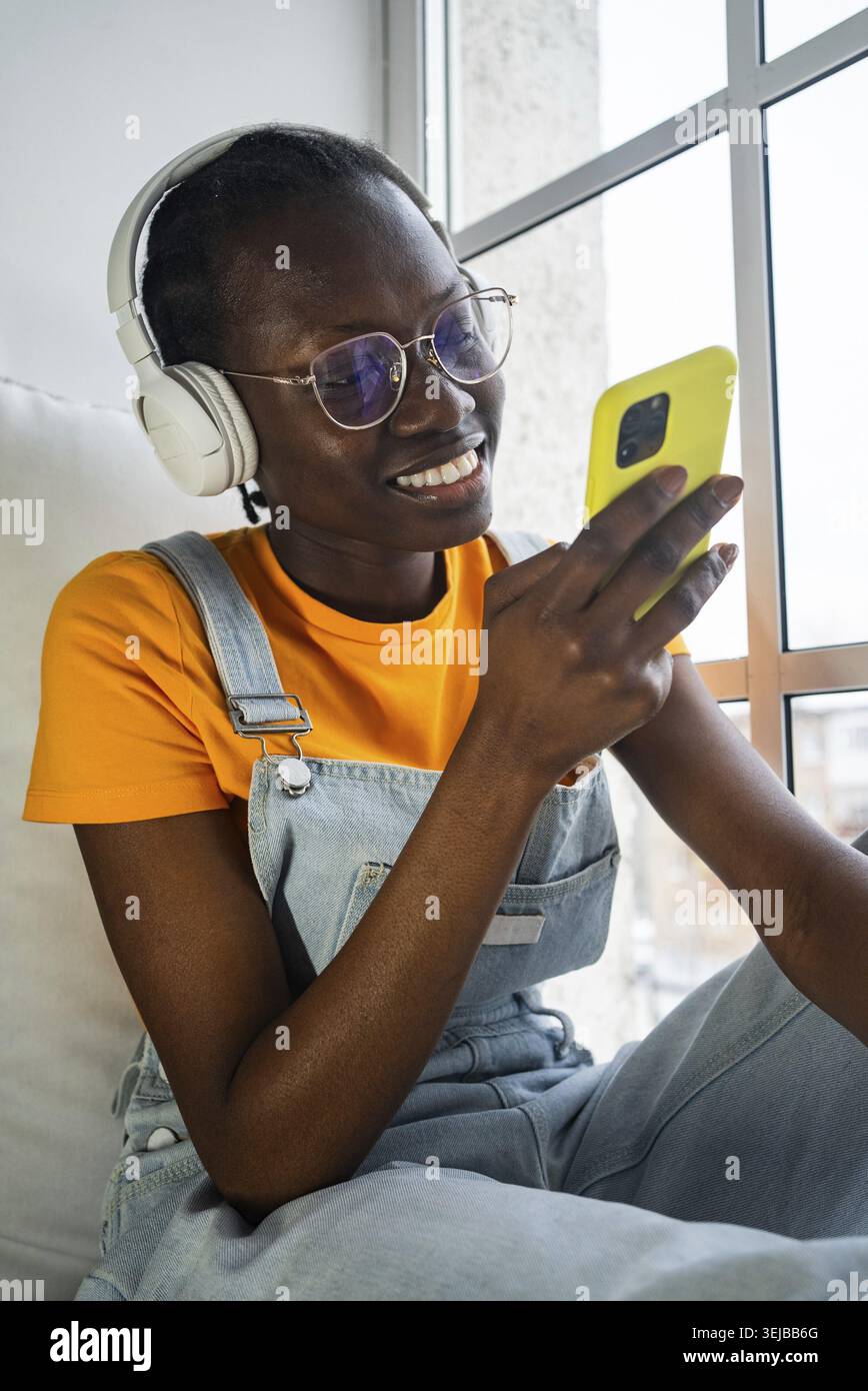 Femme afro-américaine portant des écouteurs et des lunettes, souriant tout en utilisant un smartphone. Elle est assise confortablement près d'une fenêtre, habillée décontractée, enjo Banque D'Images