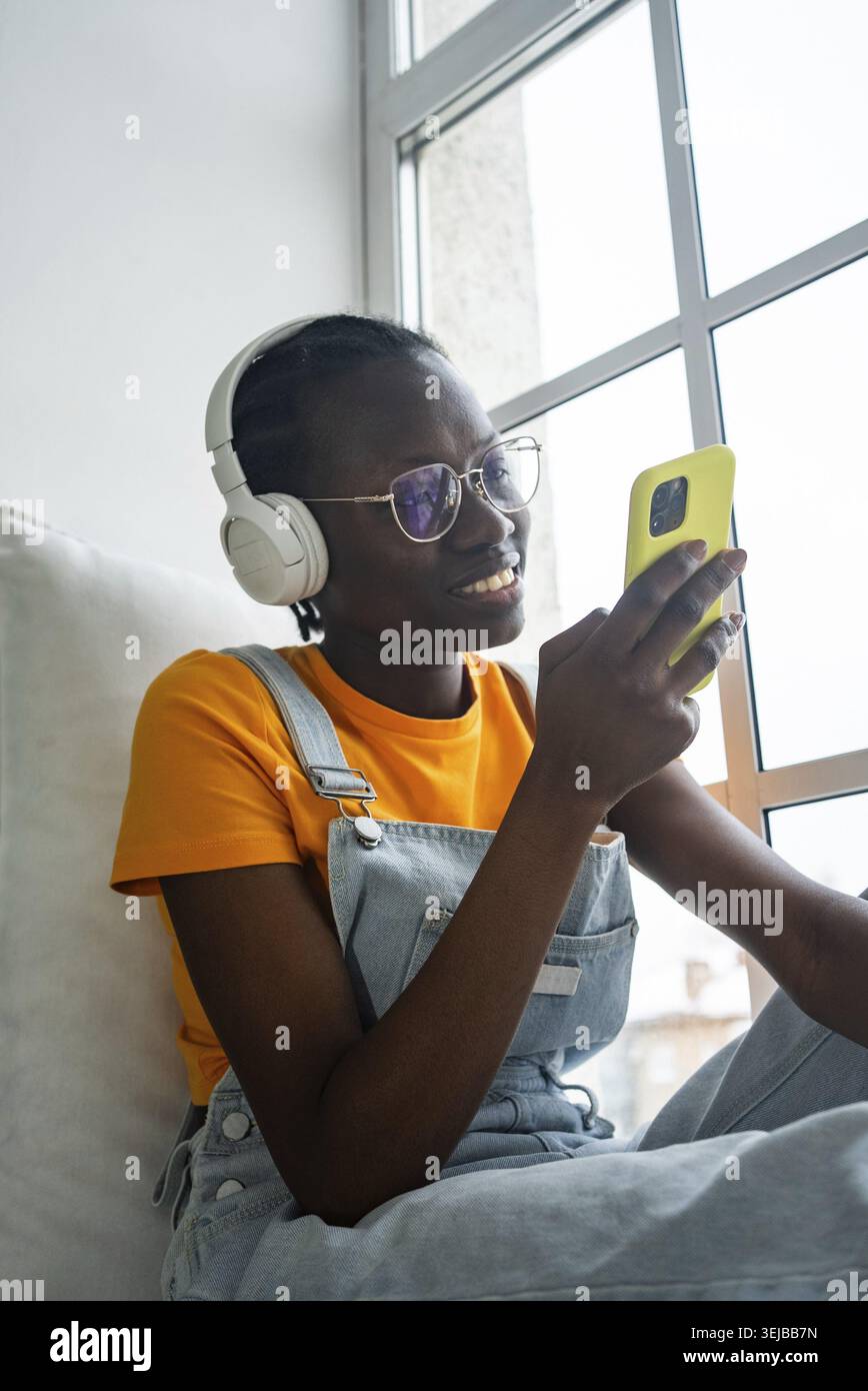 Femme afro-américaine appréciant les temps morts, portant des écouteurs et utilisant un smartphone. Assise près d'une fenêtre, elle semble détendue et satisfaite, embrassant un Banque D'Images