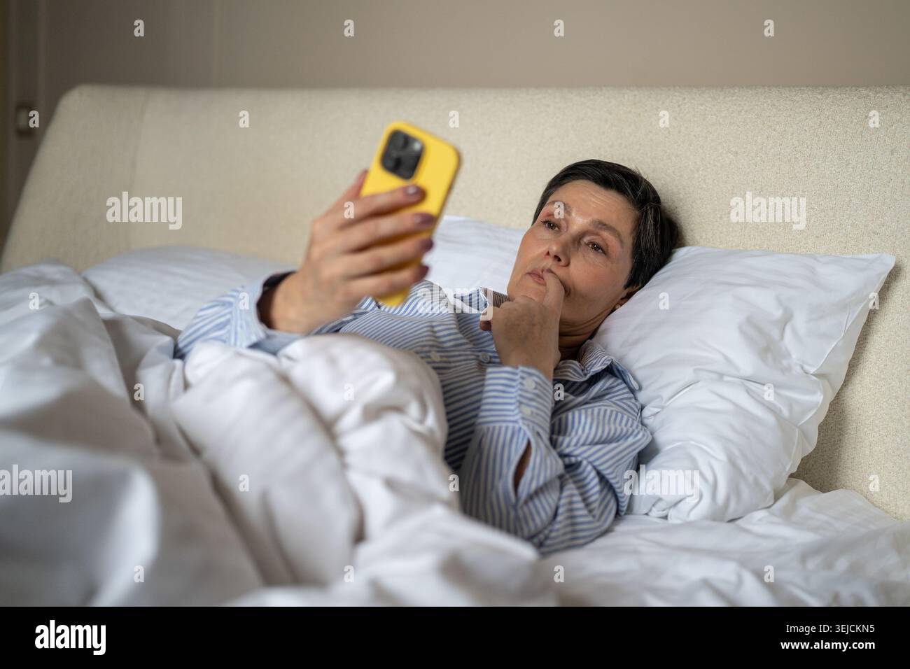 Femme âgée réfléchie dans le lit procrastinant regarder les nouvelles en ligne sur smartphone après le réveil. Banque D'Images