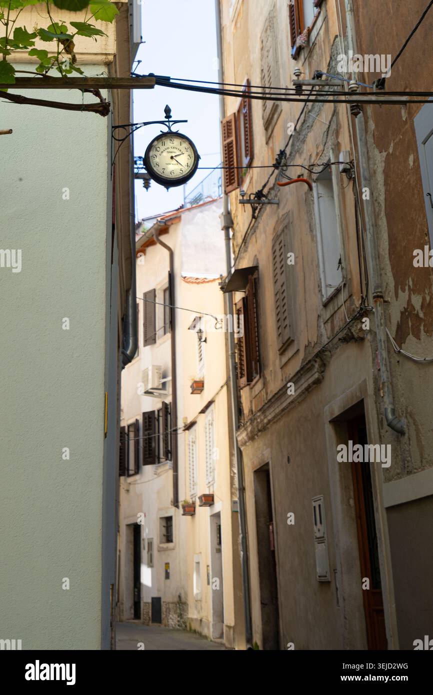 Piran, Slovénie. 26 mai 2026. Une horloge vintage est suspendue au-dessus d'une rue étroite bordée de vieux bâtiments. La lumière du soleil illumine les façades et les moulages vieillissants Banque D'Images