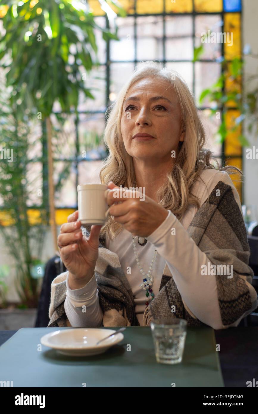 Femme mature pensive tenant une tasse de café dans un éclairage tamisé Banque D'Images