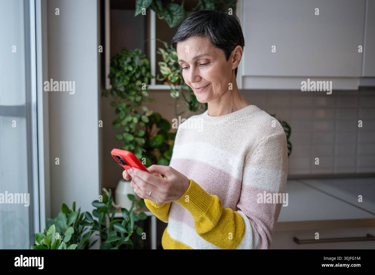 Contenu heureuse femme mature joyeuse sourit heureux, surfant sur Internet chat smartphone dans la cuisine Banque D'Images