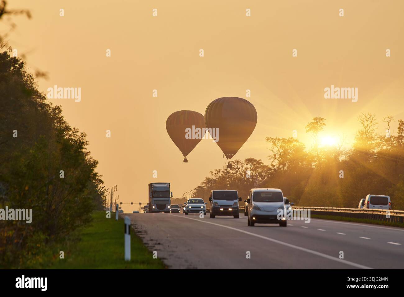 Deux montgolfières silhouettées contre un ciel brumeux de lever ou coucher de soleil, flottant au-dessus d'une autoroute achalandée avec des voitures et des camions. Des arbres bordent le bord de la route. Banque D'Images