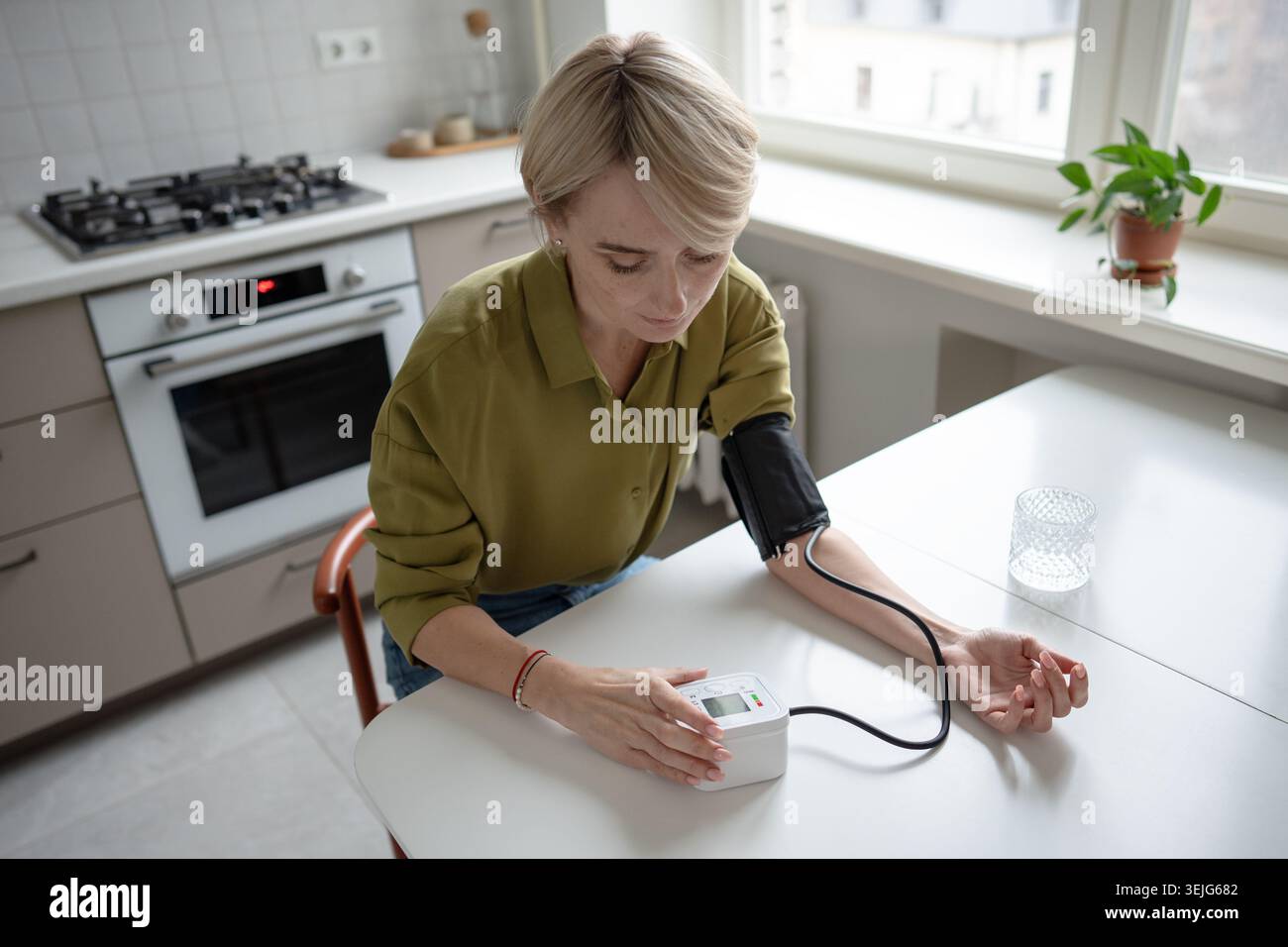 Femme d'âge moyen assise à table mesurant la pression artérielle avec tonomètre, problèmes de santé saisonniers. Banque D'Images