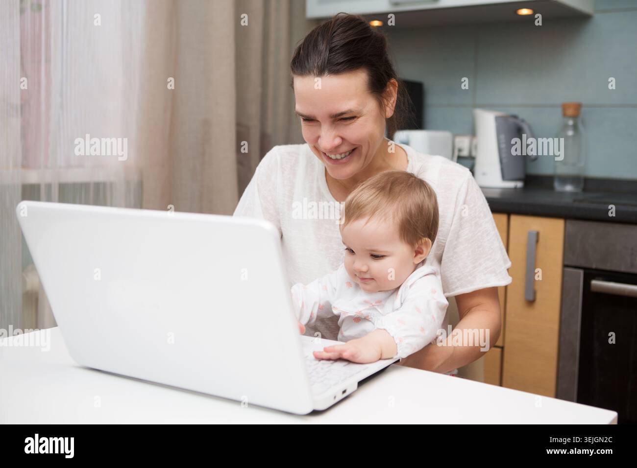 Maman et bébé. une jeune mère travaillant avec un ordinateur portable dans la cuisine Banque D'Images
