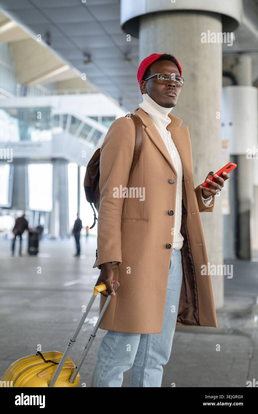 Homme de voyage afro-américain en manteau beige, chapeau rouge avec valise jaune tenant le téléphone portable, en attente des transports en commun dans ai Banque D'Images
