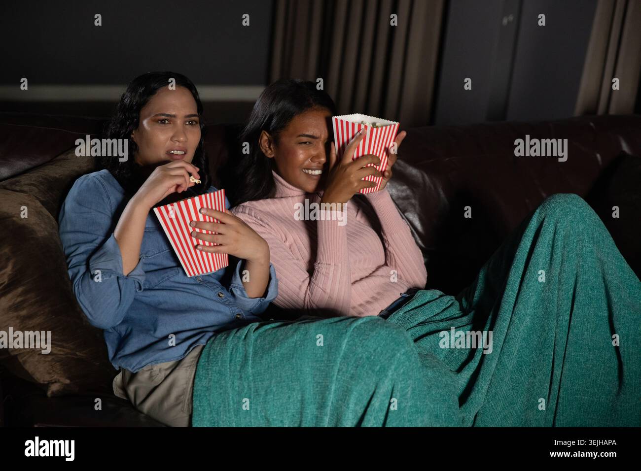 Diverses amies féminines regardant un film de suspense sur un canapé sous une couverture verte tenant des seaux de pop-corn. Confort, amitié, détente Banque D'Images