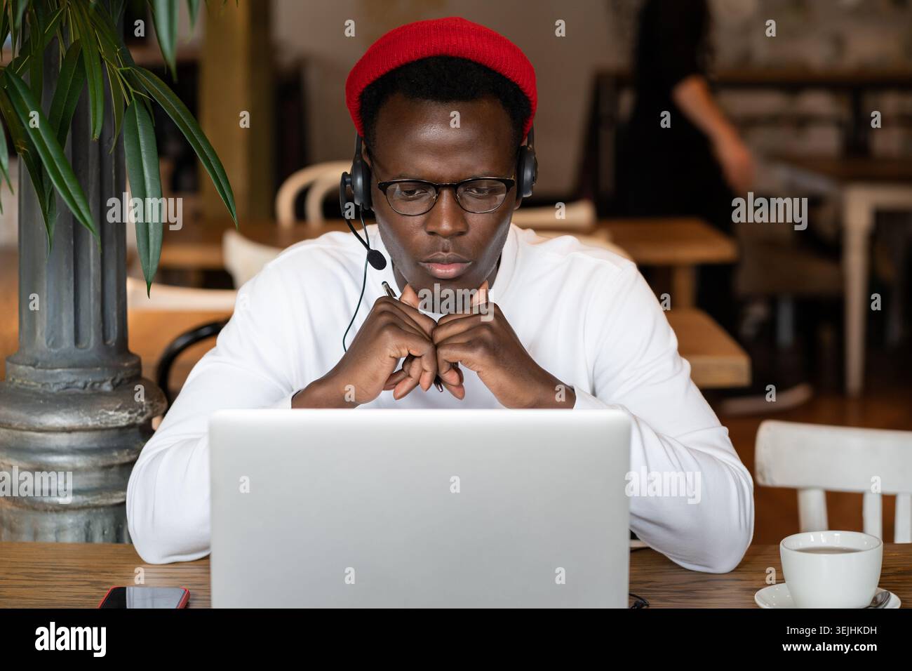 Homme noir millénaire concentré dans les écouteurs porter un chapeau rouge, regarder le webinaire éducatif sur ordinateur portable, travailler à distance en ligne dans un café. A Banque D'Images