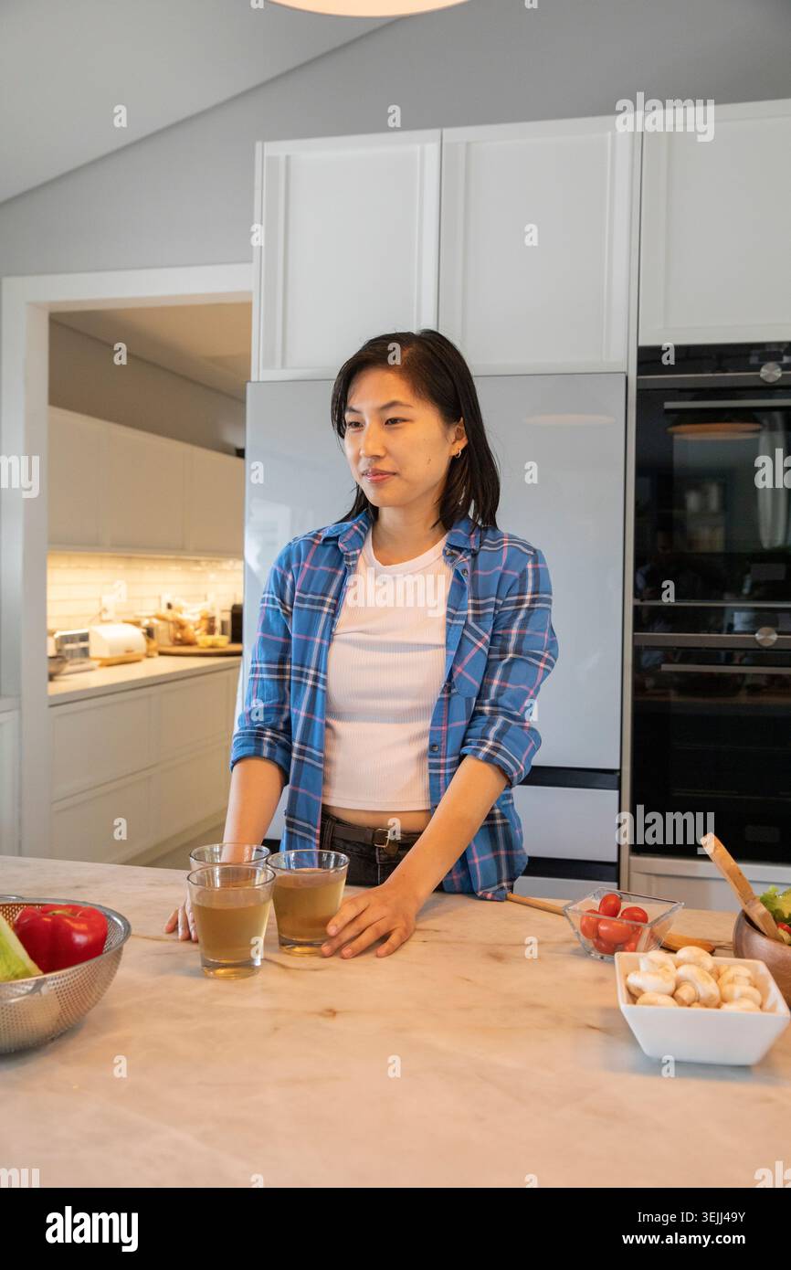 Femme asiatique tenant deux verres de jus tout en se tenant debout à l'îlot de cuisine avec des légumes frais. Moderne, minimaliste, vibrant, nous Banque D'Images