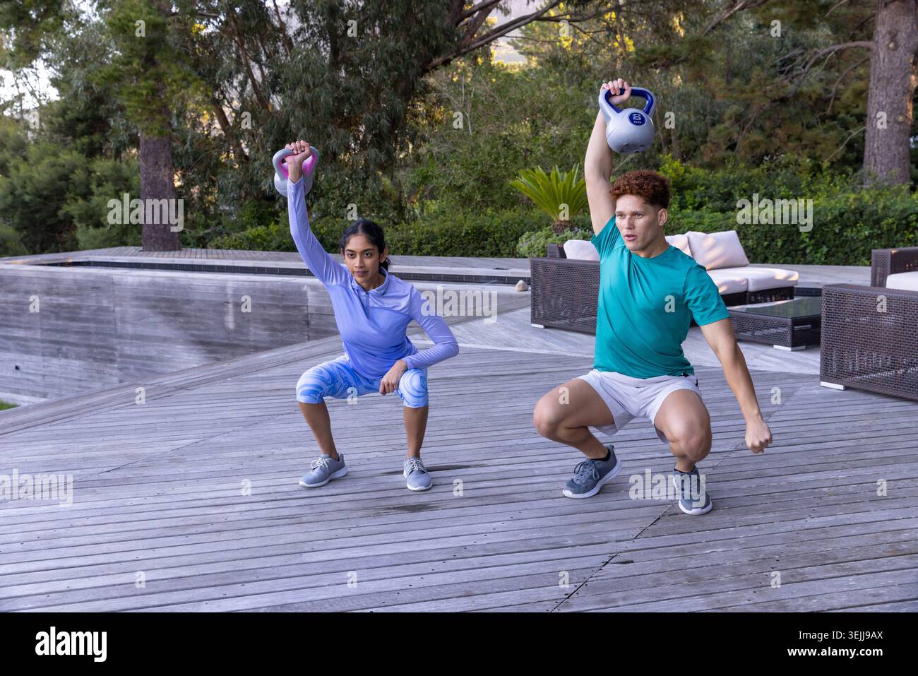 Divers partenaires d'entraînement assis en position squat sur une terrasse en bois à côté de la piscine avec kettlebells au-dessus de leurs têtes. F Banque D'Images