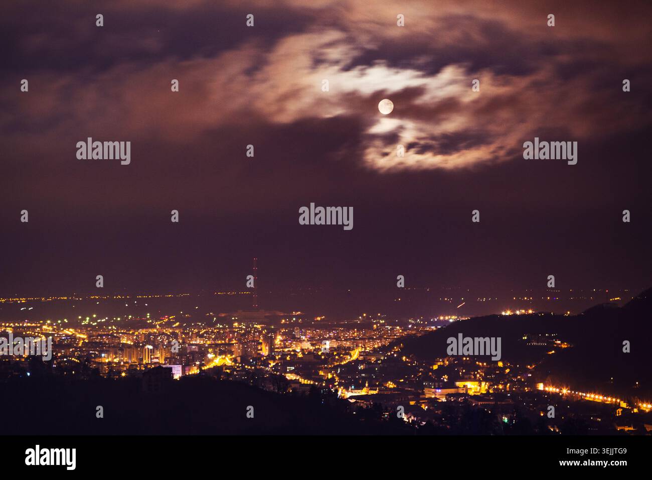 Lumières de la ville illuminant l'horizon urbain sous un ciel nuageux nocturne avec une pleine lune Banque D'Images