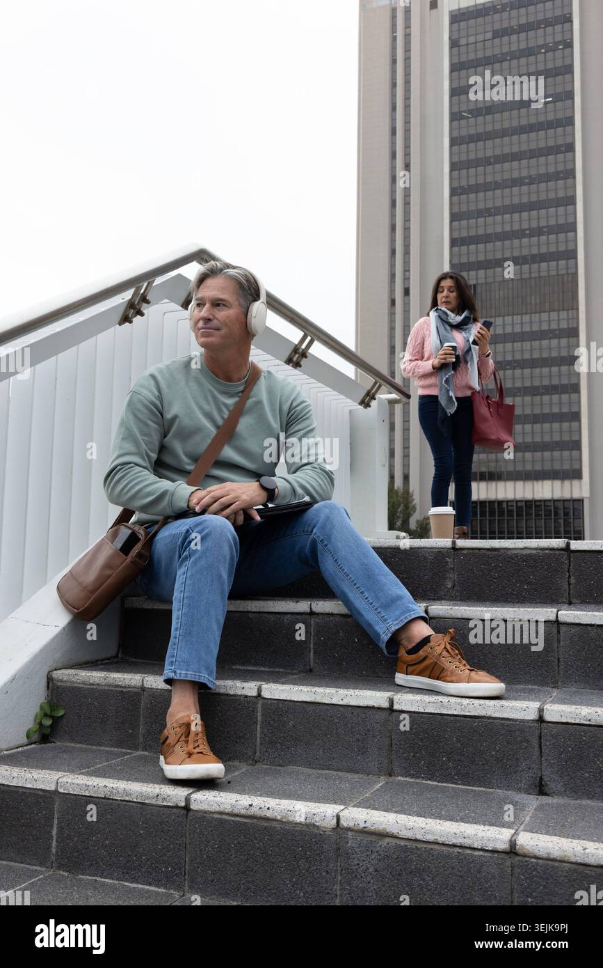 Navetteurs assis sur les marches près de la tour de bureaux, homme portant un casque utilisant une tablette, femme tenant une tasse. Urbain, escalier, architecture, Banque D'Images
