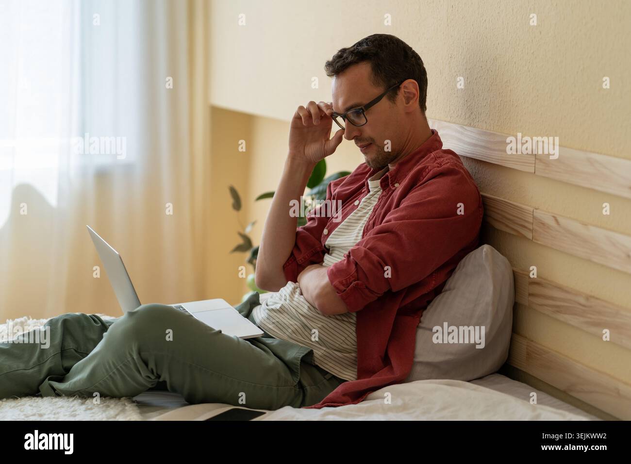 Un homme caucasien concentré est assis sur le lit dans la chambre d'hôtel pour lire les nouvelles via Internet ou consulter ses e-mails Banque D'Images