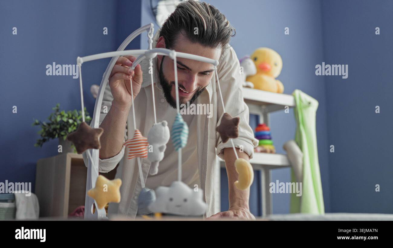L'homme se penche sur le berceau ajustant un mobile suspendu tout en souriant dans une pépinière avec des jouets en peluche, un canard en caoutchouc et des anneaux empilables ; tendresse de liaison. Banque D'Images