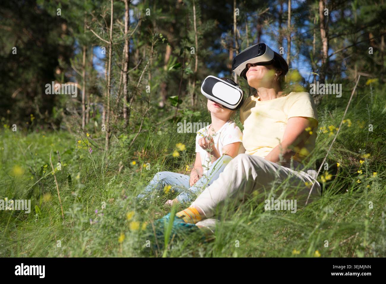 Maman et fille d'enfant jouant au jeu dans des lunettes de réalité virtuelle et détendu assis sur l'herbe dans la forêt à l'extérieur. copier Banque D'Images