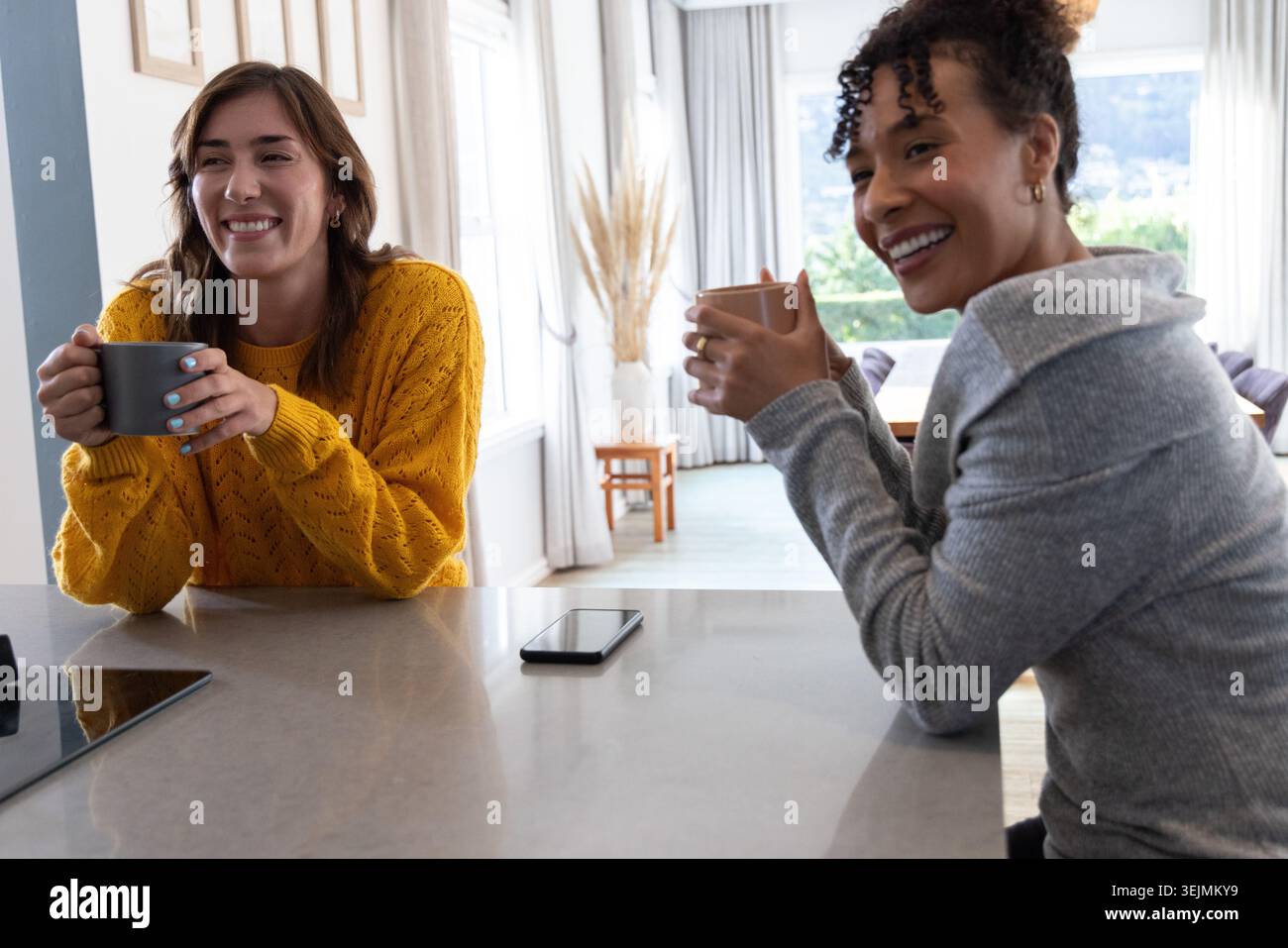 Diverses amies femmes sirotant des tasses et discutant à l'îlot de cuisine, smartphone entre eux. Comptoir, femme, ensoleillé, din Banque D'Images