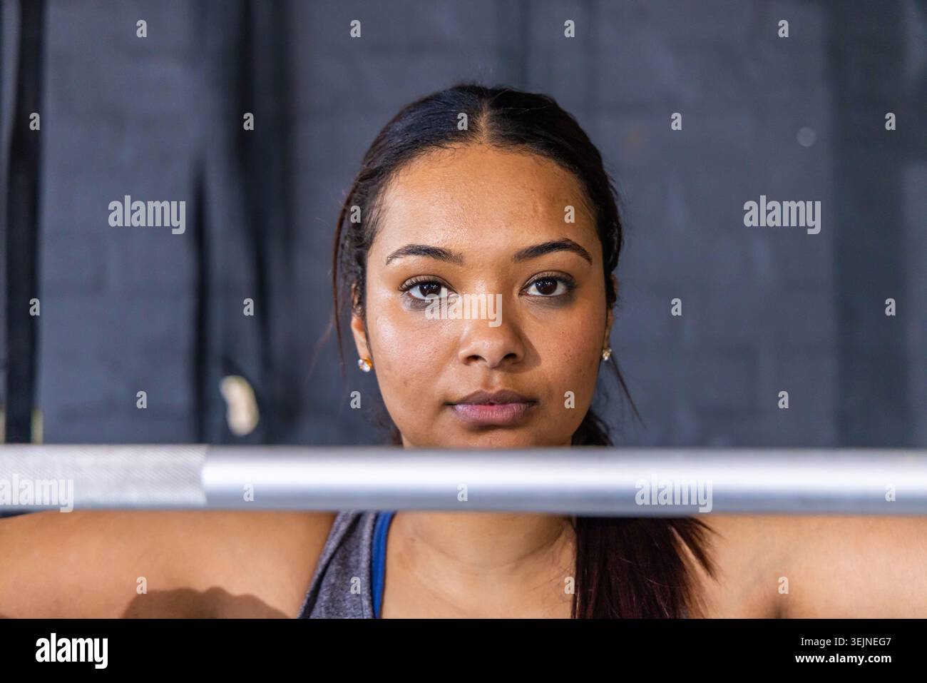 Femme debout derrière une barre horizontale au gymnase, portant un débardeur gris et une sangle bleue. Force, concentration, athlétisme, forme physique, tr Banque D'Images