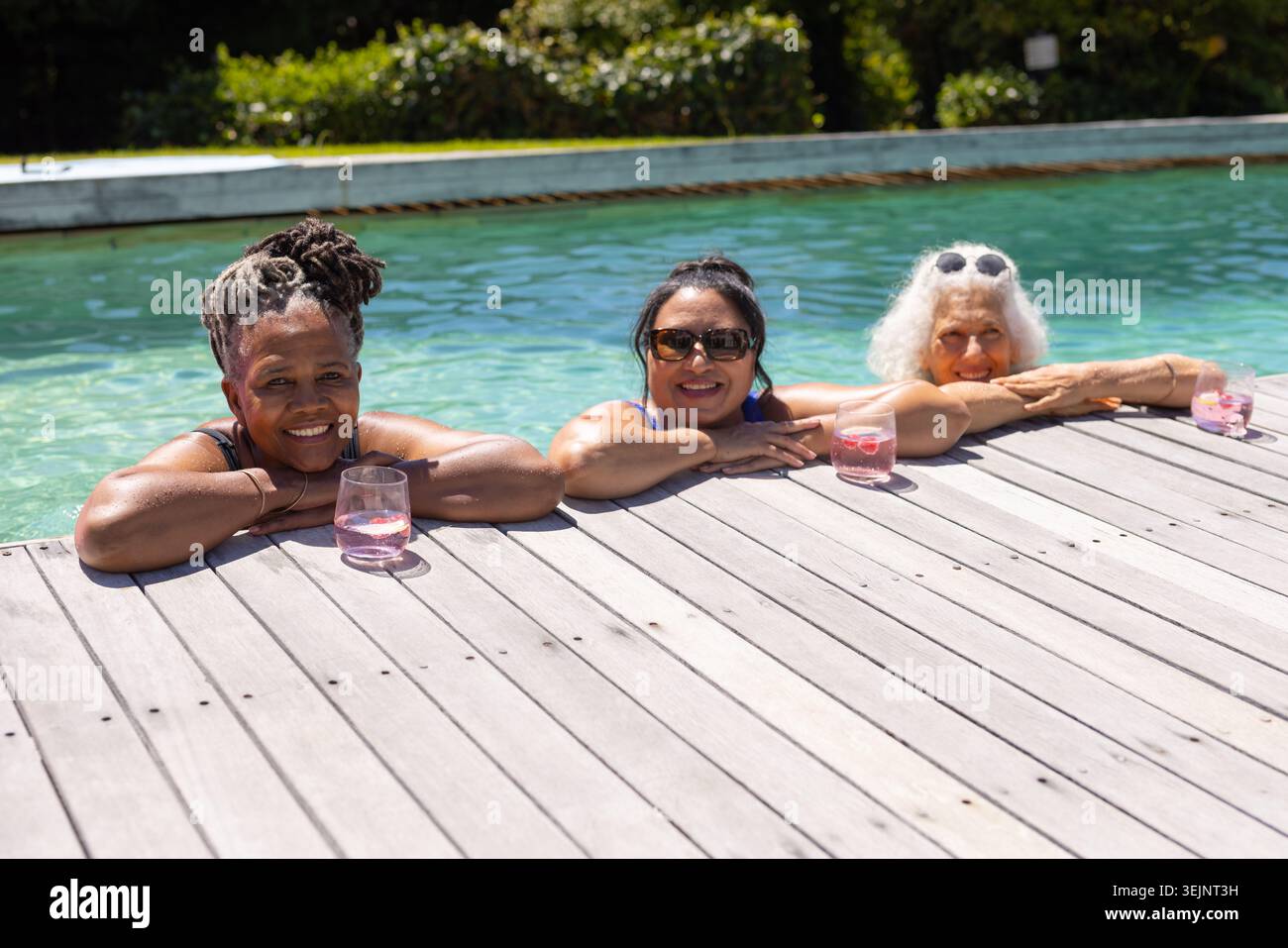 Diverses amies féminines réchauffant la peau au soleil, appuyées sur la terrasse en bois à la piscine, sirotant des boissons roses. Piscine, trio, lunettes de soleil, turquo Banque D'Images