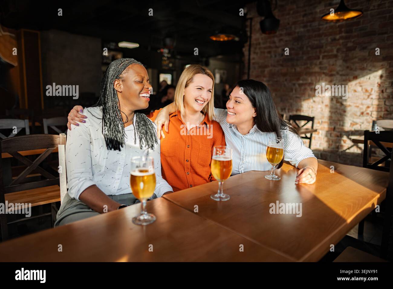 Trois amies diverses dégustant de la bière et riant dans un pub Banque D'Images