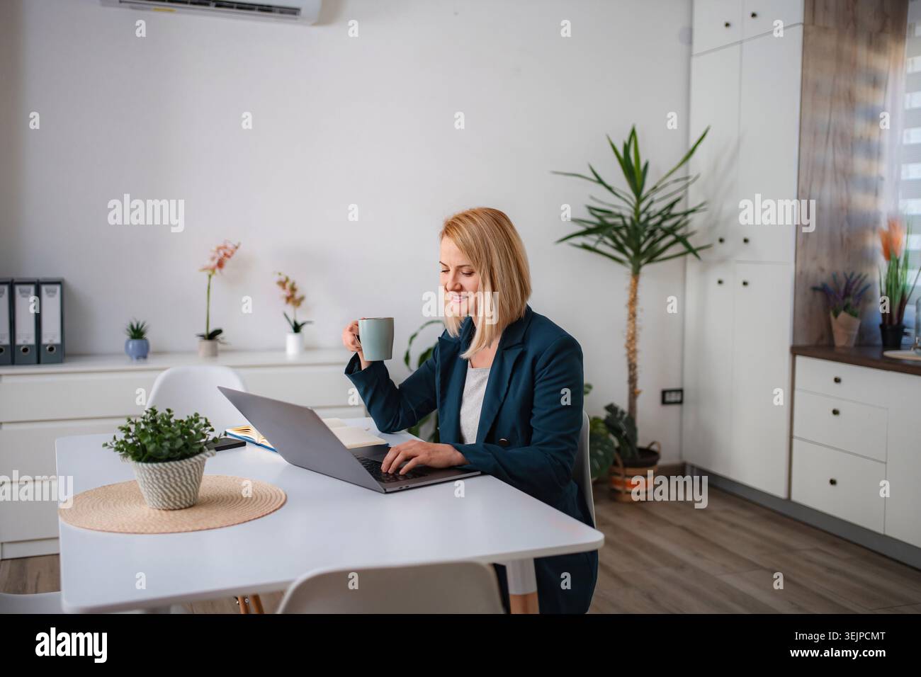 Femme souriante travaillant à la maison à l'aide d'un ordinateur portable et buvant du café Banque D'Images