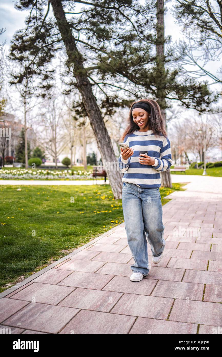 Jeune femme noire marchant dans le parc vérifiant smartphone Banque D'Images