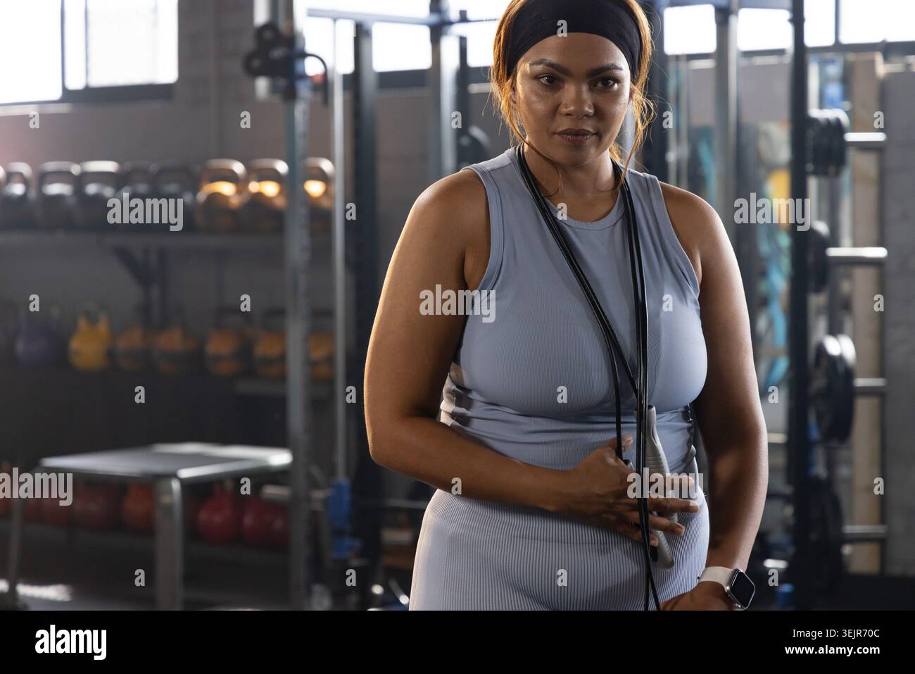 Femme afro-américaine debout dans un gymnase commercial portant des vêtements d'activité ajustés, corde à sauter, bandeau. Kettlebell, poids, banc, BA Banque D'Images