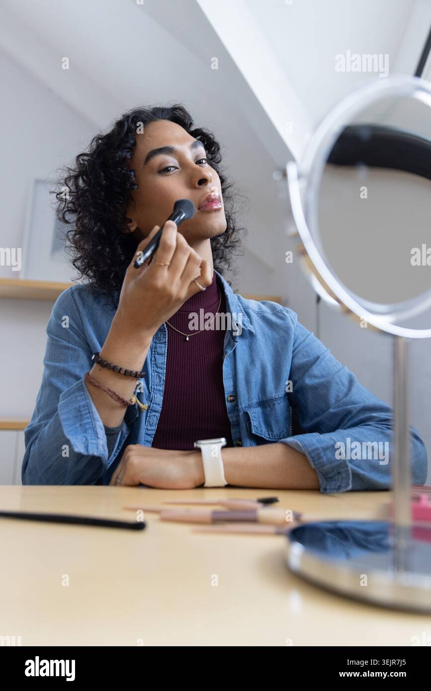 Personne non binaire appliquant le maquillage avec la brosse à la table de vanité en utilisant le miroir rond portant le dessus en denim. Cosmétiques, beauté, autosoins, Banque D'Images