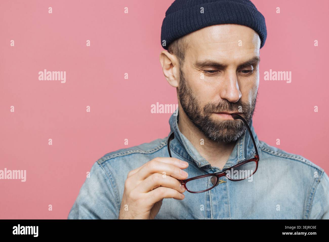 Homme d'âge moyen tenant des lunettes sur fond rose portant chemise en denim et bonnet en maille Banque D'Images