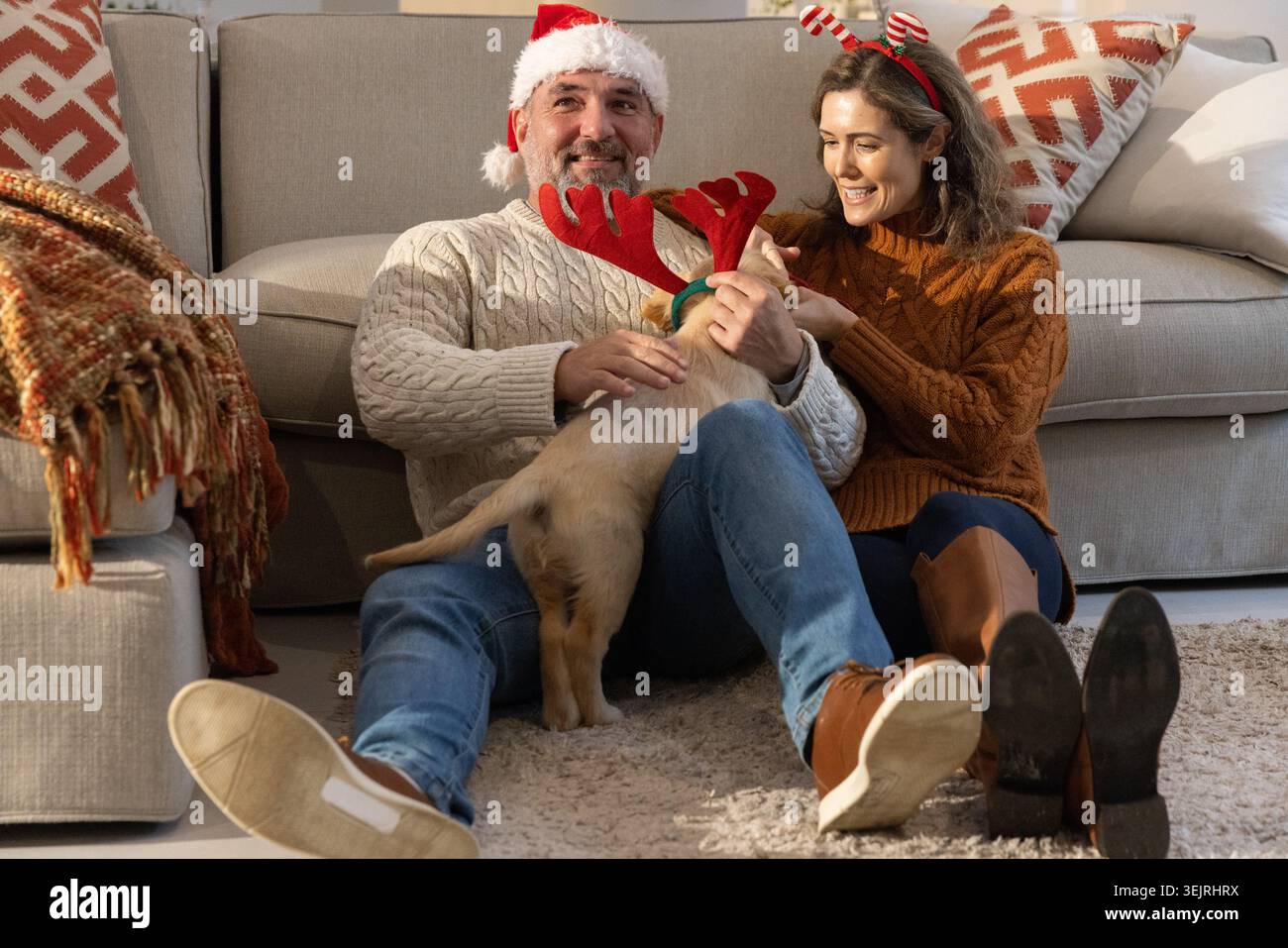 Couple assis sur un tapis en peluche dans le salon caressant chiot portant des bois rouges et chapeau de Père Noël. Confortable, maison, duo, chien, festif, dayli Banque D'Images
