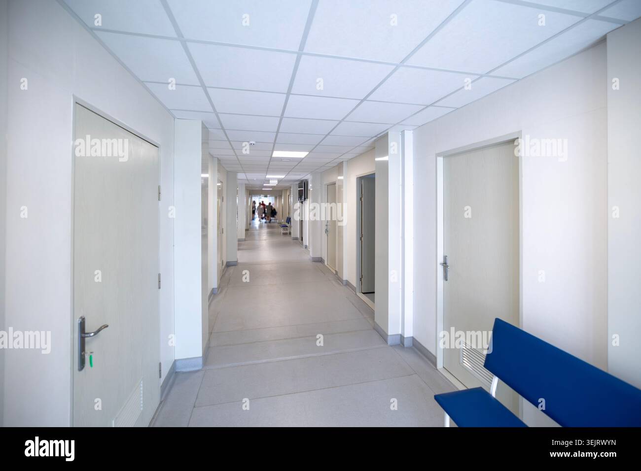 Un couloir d'hôpital intérieur long et lumineux avec des murs peints en blanc, des portes en bois fermées, un plafond suspendu avec des lumières carrées, un banc bleu sur la R Banque D'Images