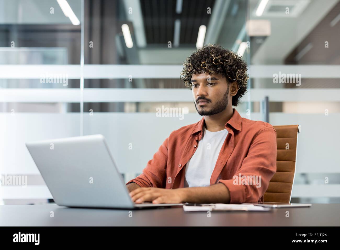 Jeune homme indien aux cheveux bouclés tapant intentionnellement sur un ordinateur portable, se concentrant sur son travail à un bureau dans un espace de travail de co-working contemporain avec des cloisons en verre Banque D'Images
