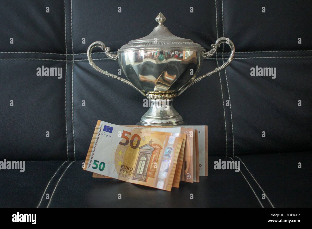 Photo conceptuelle d'un trophée de championnat en argent brillant placé derrière une pile de billets de 50 euros. Ensemble contre une surface en cuir noir foncé, symboli Banque D'Images