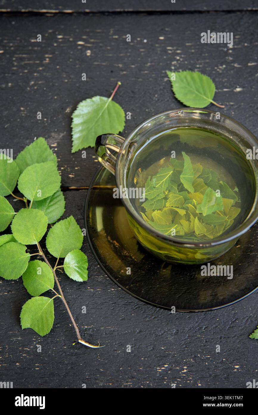 Au-dessus de la vue du thé de feuille d'arbre de bouleau dans une tasse en verre, Betula tisane médicinale dans une tasse claire. Fond de planche de bois de couleur noire, feuilles et branche. Banque D'Images
