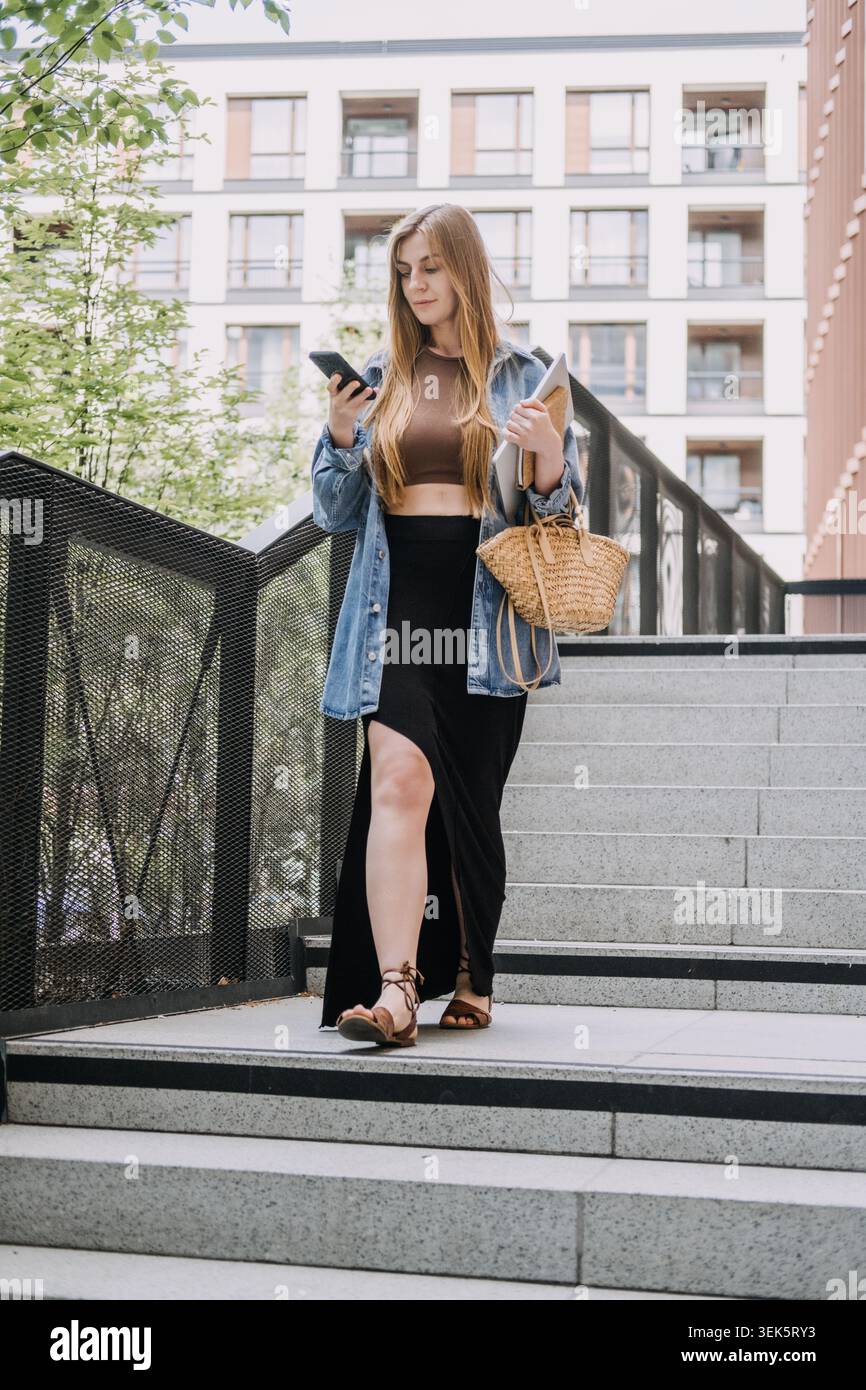 Jeune femme se tient sur les escaliers de la ville regardant smartphone tout en tenant un ordinateur portable. Prise de décision à la volée, vie professionnelle active, affaires Banque D'Images