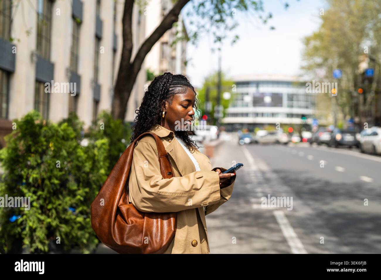 Femme afro-américaine réfléchie debout dans la rue attendant les transports en commun et vérifiant l'horaire au téléphone, femme noire sérieuse attendant le bus à la gare en ville et défilant sur smartphone. Banque D'Images