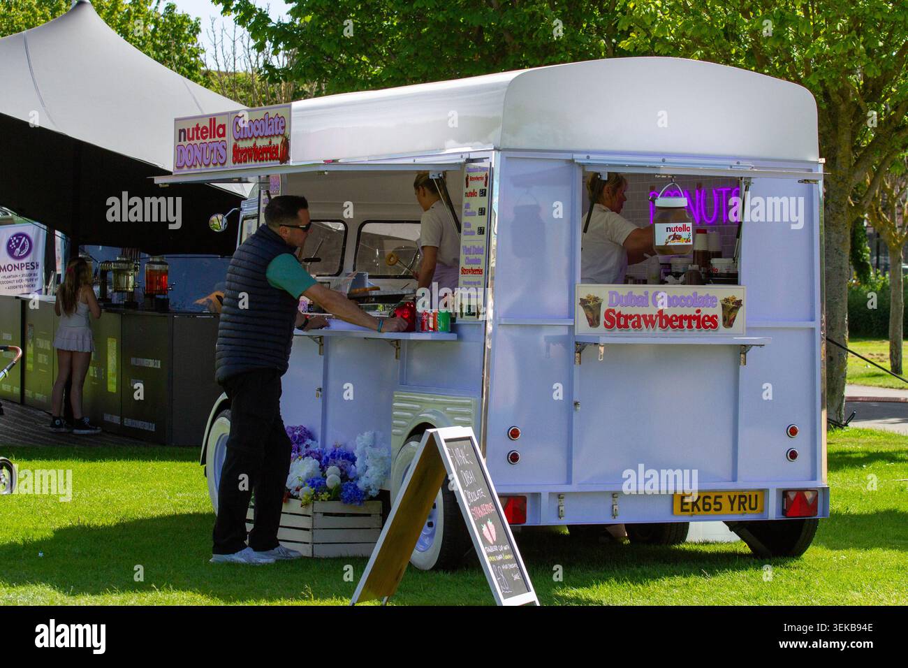 Un client se tient à la fenêtre de service d'une camionnette de dessert mobile de style rétro blanche faisant la publicité de beignets à la Nutella et de fraises au chocolat. Banque D'Images