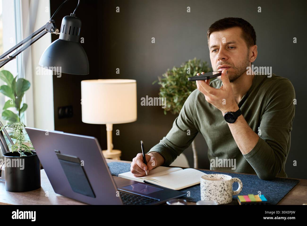 Homme parlant au smartphone et écrivant des notes au bureau à la maison avec ordinateur portable, tasse à café et lumière intérieure chaude. Concept d'assistant vocal, de planification quotidienne, de travail à distance et de technologie d'IA quotidienne. Banque D'Images