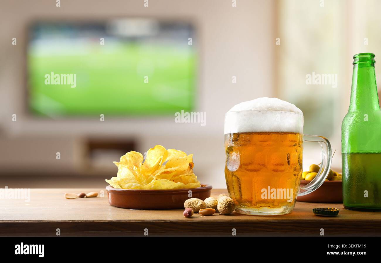 Tasse de bière froide avec des chips et des collations sur une table en bois tandis qu'un match de football joue floue en arrière-plan à la maison. Banque D'Images