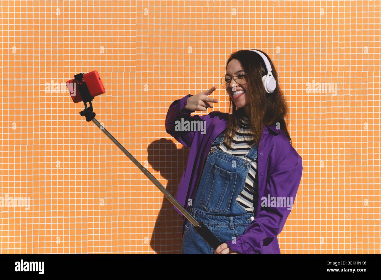 Jeune femme souriante, portant des écouteurs et des lunettes, prenant un selfie à l'extérieur avec un mur orange Banque D'Images