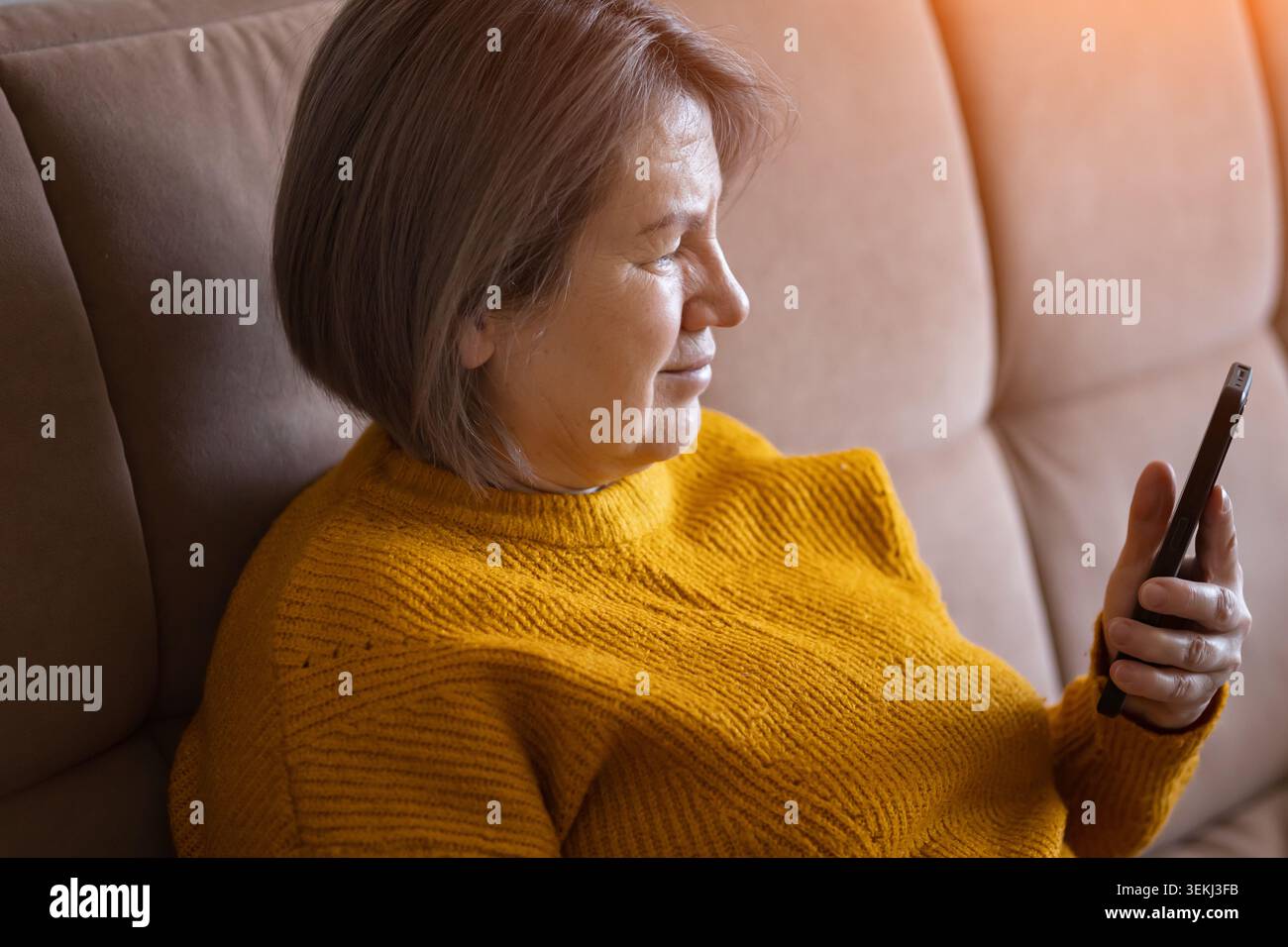 Femme âgée se relaxant sur un canapé et en utilisant un smartphone dans un salon chaleureux et confortable par la lumière de la fenêtre Banque D'Images